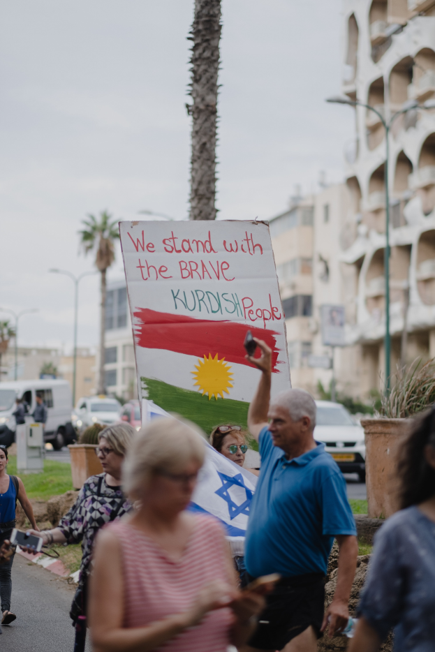 Eine Gruppe von Menschen marschiert nachts durch eine Straße und hält ein Schild mit der Aufschrift "Wir stehen zu den tapferen kurdischen Menschen", mit einem geparkten Van, Straßenlaternen, Bäumen, Gebäuden und einem klaren blauen Himmel im Hintergrund.