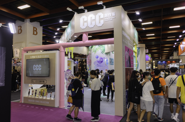 A group of people at a trade show booth labeled "ccc creative comic collection," with text boards, ceiling lights, and a screen on the left.