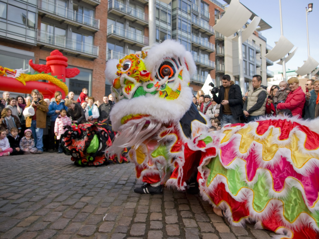 Ein farbenfrohes chinesisches Neujahrsfest in Amsterdam mit einem Löwen tanzen im Vordergrund und einer Menge Schaulustiger, einige halten Kameras, vor einem Hintergrund aus Gebäuden, Laternenmasten und einem klaren blauen Himmel.