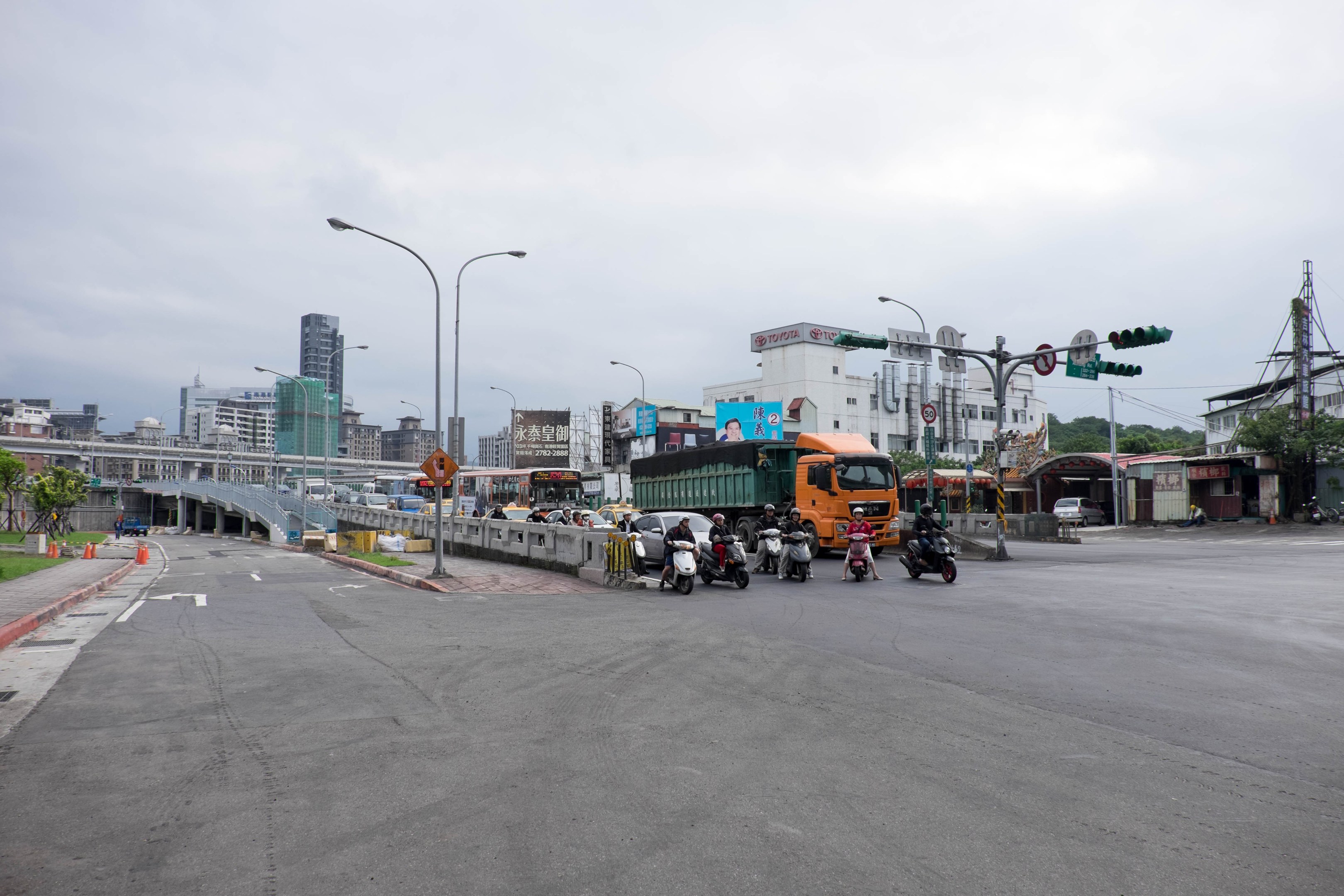 A busy city street with multiple vehicles, including a truck and a car, lined with light poles, traffic signals, signboards, a bridge, trees, buildings, and a cloudy sky.
