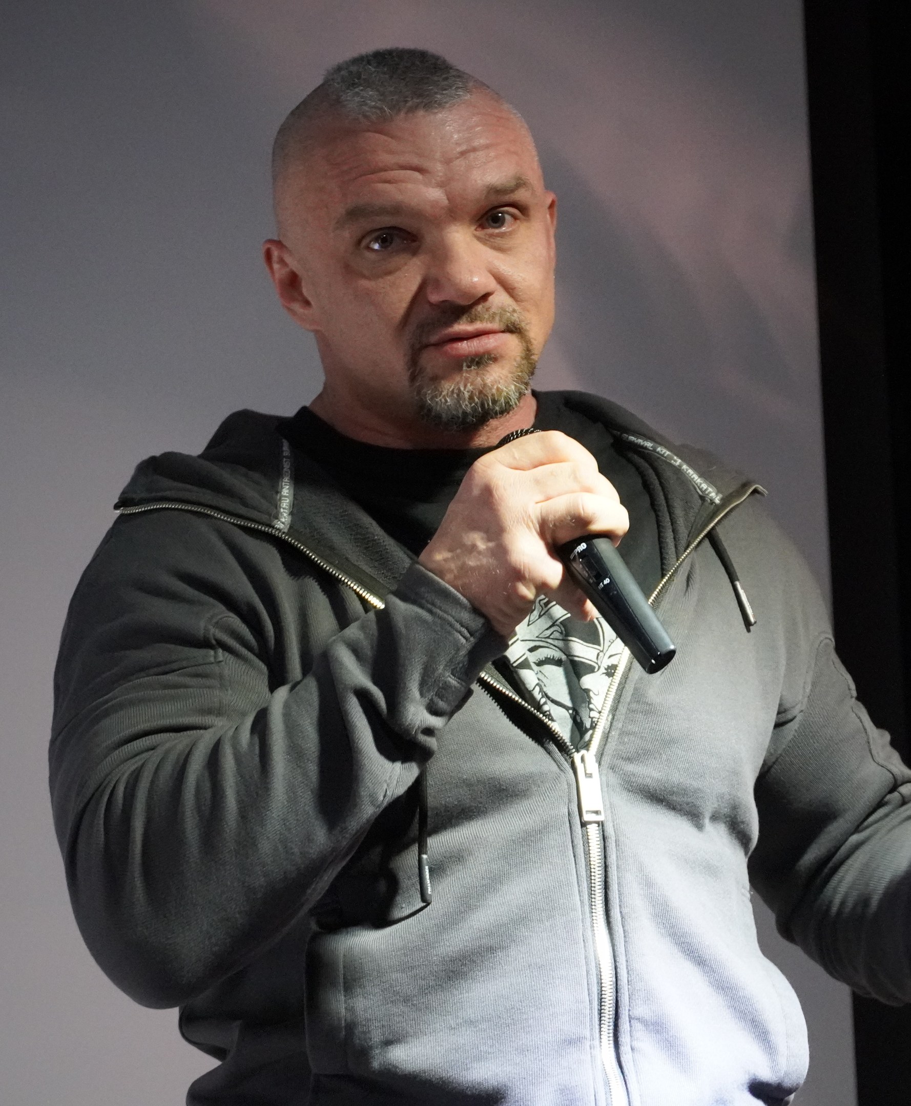 Ein Mann in einer schwarzen Kapuzenjacke und einem schwarzen Anzug mit einem weißen Hemd, der ein Mikrofon hält und selbstbewusst vor einer Wand steht.