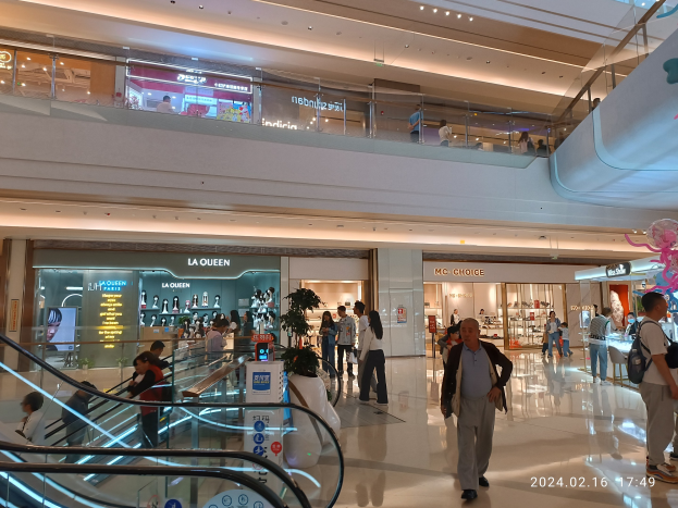 Interior of a shopping mall with people walking, escalators, illuminated ceiling lights, store signs, informational boards, potted plants, and a visible watermark noting the mall's recent reopening post-coronavirus pandemic.
