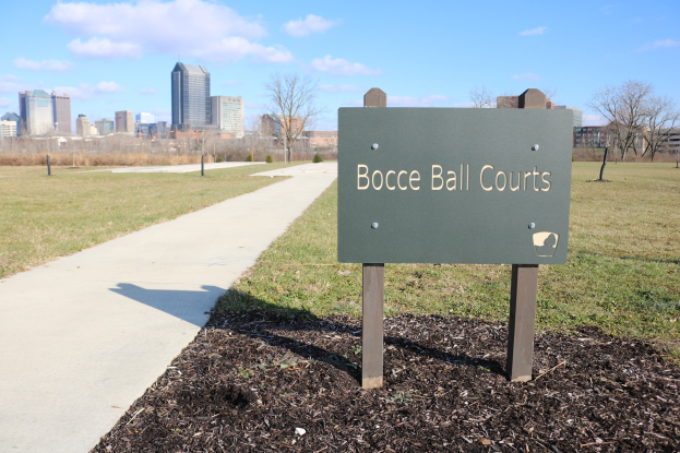Ein Schild am Boden trägt die Aufschrift "Boccia-Plätze in Indianapolis, Indiana" in der Nähe eines Weges, Bäume, Pfähle, ein Zaun, Gebäude und einen bewölkten Himmel.