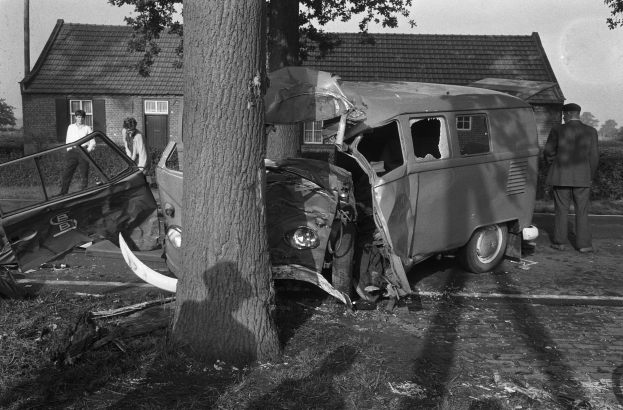 Ein Schwarz-Weiß-Bild eines verbeulten Autos am Straßenrand, mit einem Baum im Vordergrund, einigen Menschen in der Nähe und Häusern, Bäumen und einem Mast im Hintergrund.