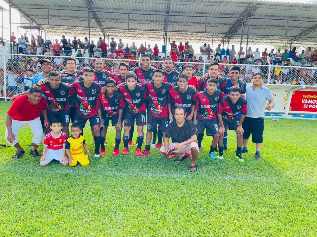 Mannergruppe feiert auf einem Fußballfeld mit einem Siegesschild, auf dem "Fotos Ugarte U-17 vs U-18" steht, unter einem bewölkten Himmel.