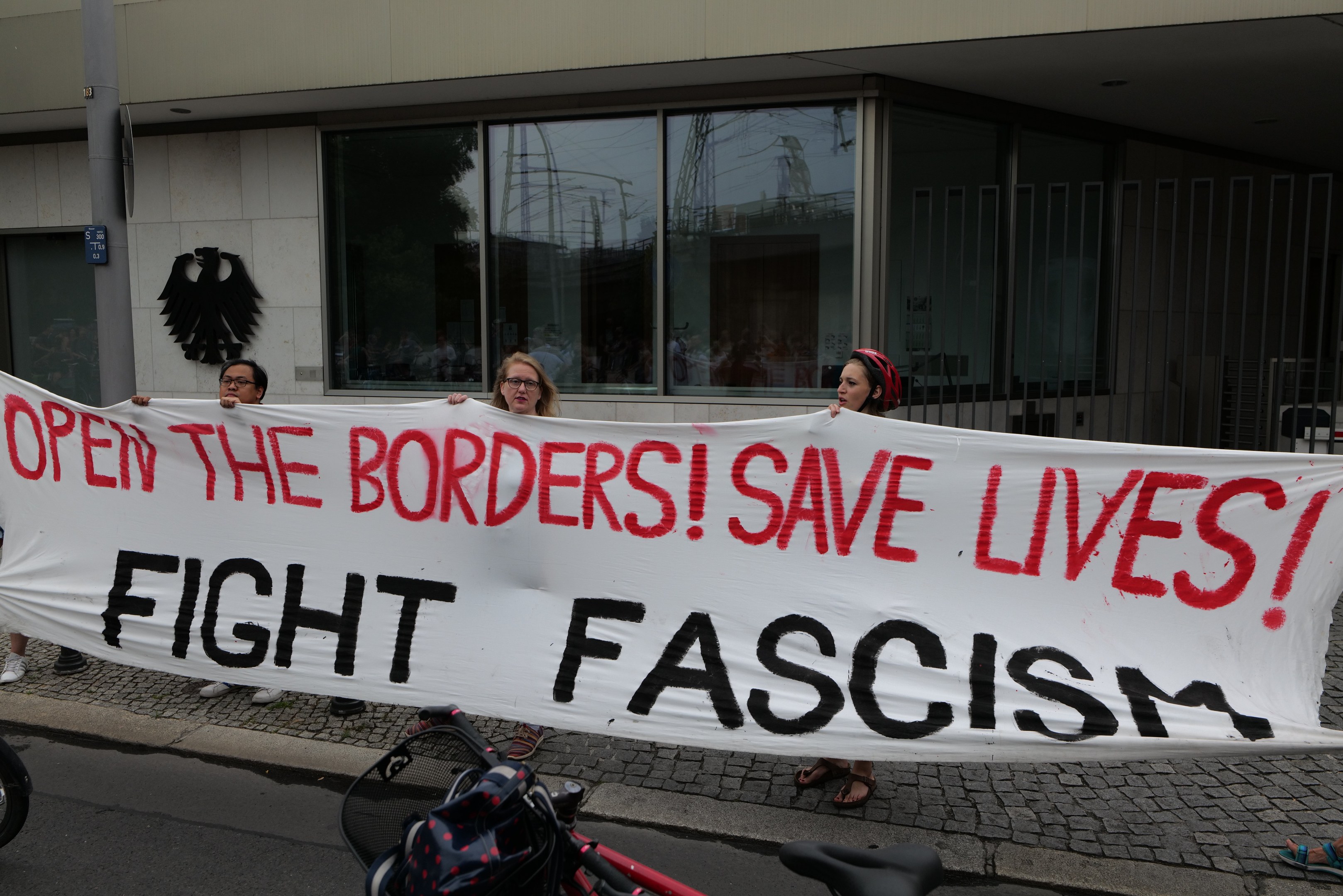 Gruppe von Menschen mit einem Banner "Öffnet die Grenzen, rettet Leben, kämpft gegen Faschismus" vor einem Gebäude mit Glasfenstern und einem Mast, mit einem Fahrrad und einer Tasche auf der Straße.