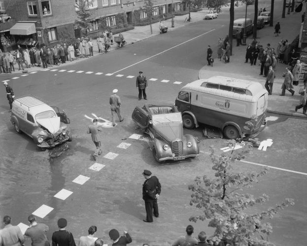 Eine Schwarz-Weiß-Szene eines Autounfalls an einer Straßenkreuzung mit mehreren Fahrzeugen, darunter ein Van, einer Gruppe von Menschen drumherum und im Hintergrund Gebäude, Bäume, Pfosten und Texttafeln.