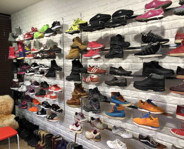 Ein Schuhgeschäftsinneres mit mehreren Ständern, die verschiedene Arten von Schuhen zeigen, einem Stuhl auf der linken Seite mit einem Teddybär darauf und einer Wand im Hintergrund.