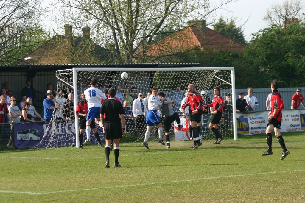 Spieler spielen Fußball auf einem Feld mit einem Tor, während Zuschauer dahinter stehen; Bäume und Häuser sind im Hintergrund sichtbar.