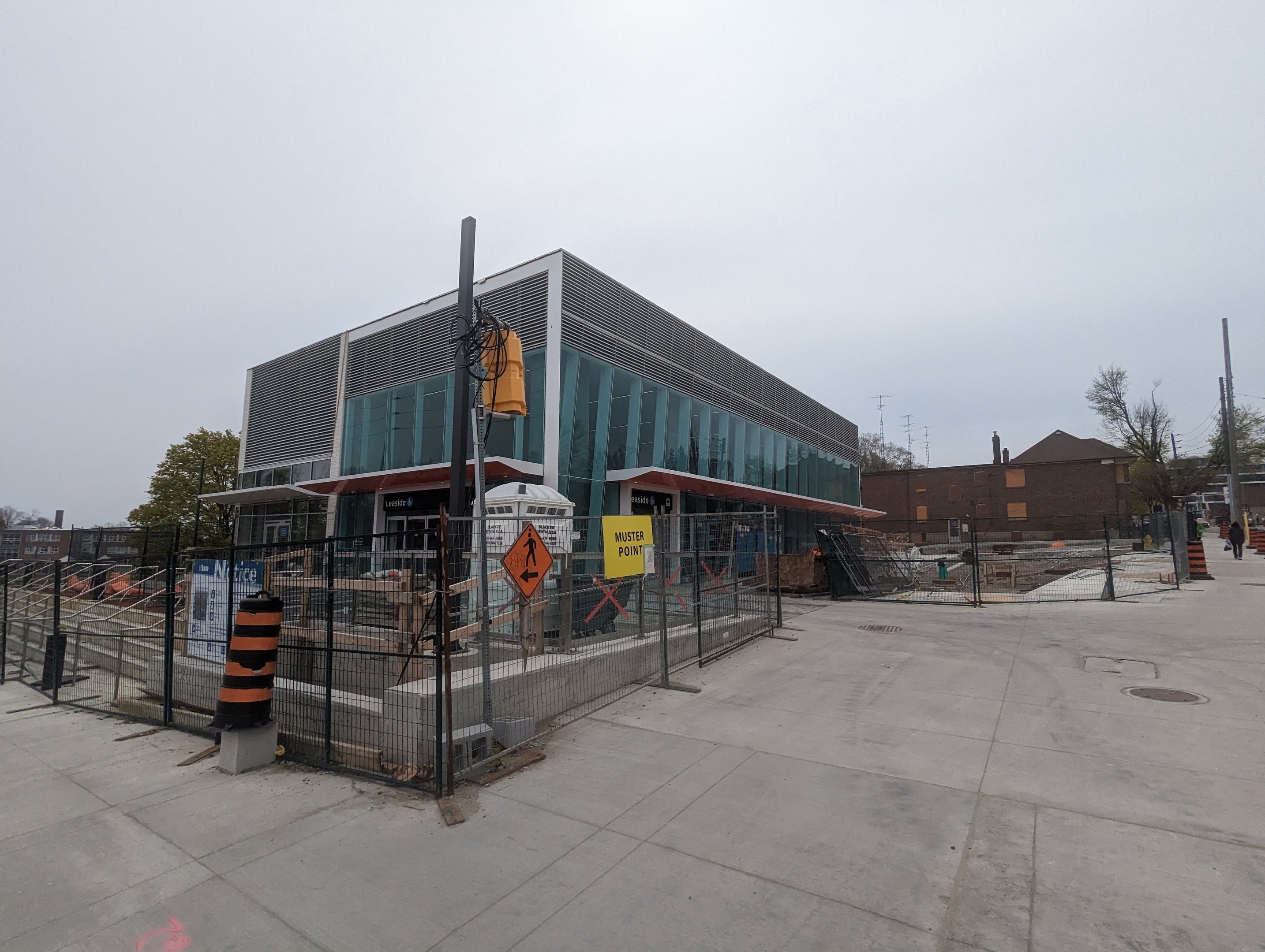 Large modern school building with many windows, surrounded by a metal fence, trees, signboards, and a few people and vehicles outside under a cloudy sky.
