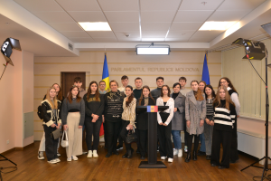 Eine Gruppe von Menschen steht vor einem Podium mit Mikrofon, umgeben von Fahnen, einer Wand mit dem Text "Moldova Youth Congress 2018", einer Tür, einem Dach mit Deckenlampen und Ständern mit Lampen.