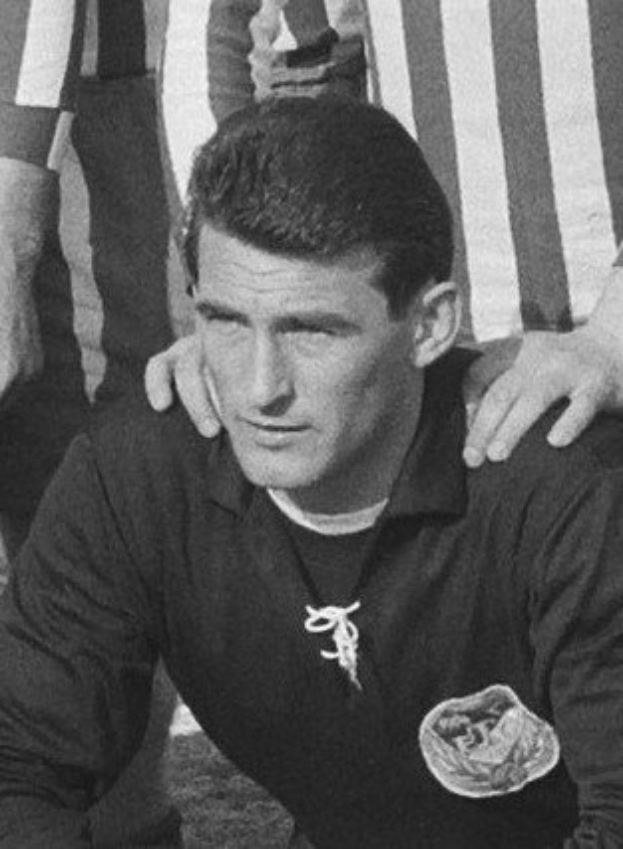 Schwarzes und weißes Foto eines Mannes, der auf einem grasbewachsenen Fussballfeld sitzend eine T-Shirt und eine Kette trägt, mit zwei Personen hinter ihm.