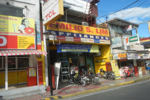 Eine belebte Straßenszene mit Gebäuden, geparkten Motorrädern, Straßenlaternen, Strommasten, Stromkabeln, Bäumen und einem bewölkten Himmel, mit einem Geschäft namens "Wilio S Lim Appliances" im Vordergrund.