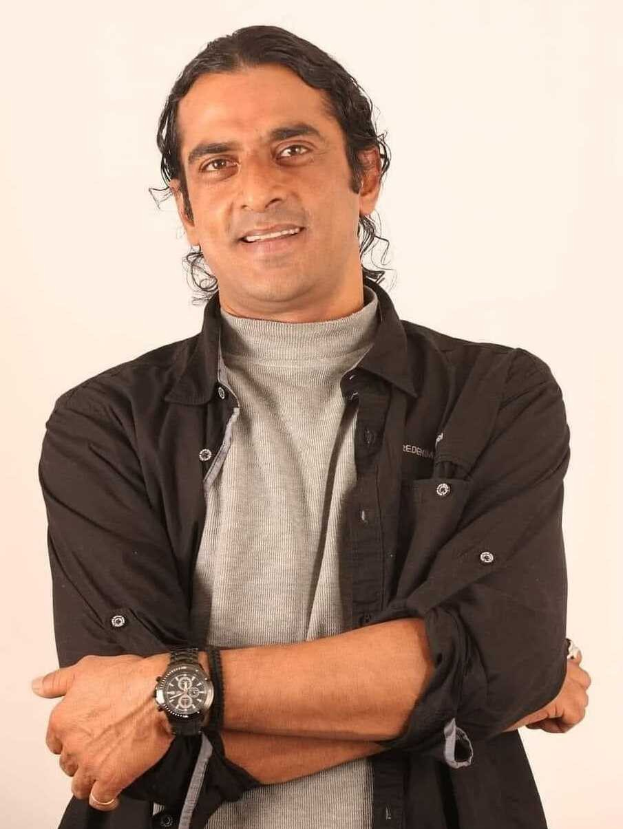 Ein Mann in einem schwarzen und aschfarbenen Kleid mit überkreuzten Armen, der eine Uhr trägt und vor einem weißen Hintergrund steht, in Gedanken versunken.