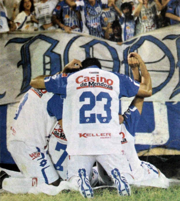 Gruppe von Fußballspielern, die auf dem Boden vor einer jubelnden Menge feiern, mit einem teilweise sichtbaren Banner im Hintergrund.