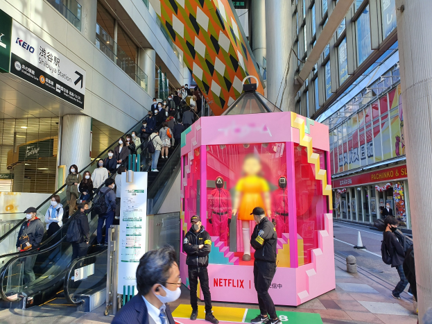 Eine Gruppe von Menschen steht vor einem Gebäude mit Glaswänden und Texttafeln, einige tragen Masken, während eine Rolltreppe mit Menschen zu sehen ist; ein pinker Kasten in der Mitte zeigt Schaufensterpuppen und trägt den Text "Netflix's new Netflix Booth in Tokyo."