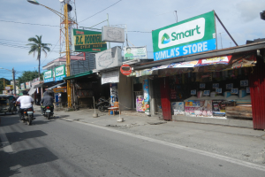 Eine belebte Straßenszene mit Menschen auf Motorrädern und Autos, umgeben von Gebäuden, Geschäften, Schildern, Strommasten, Straßenlaternen, Bäumen und einem bewölkten Himmel.