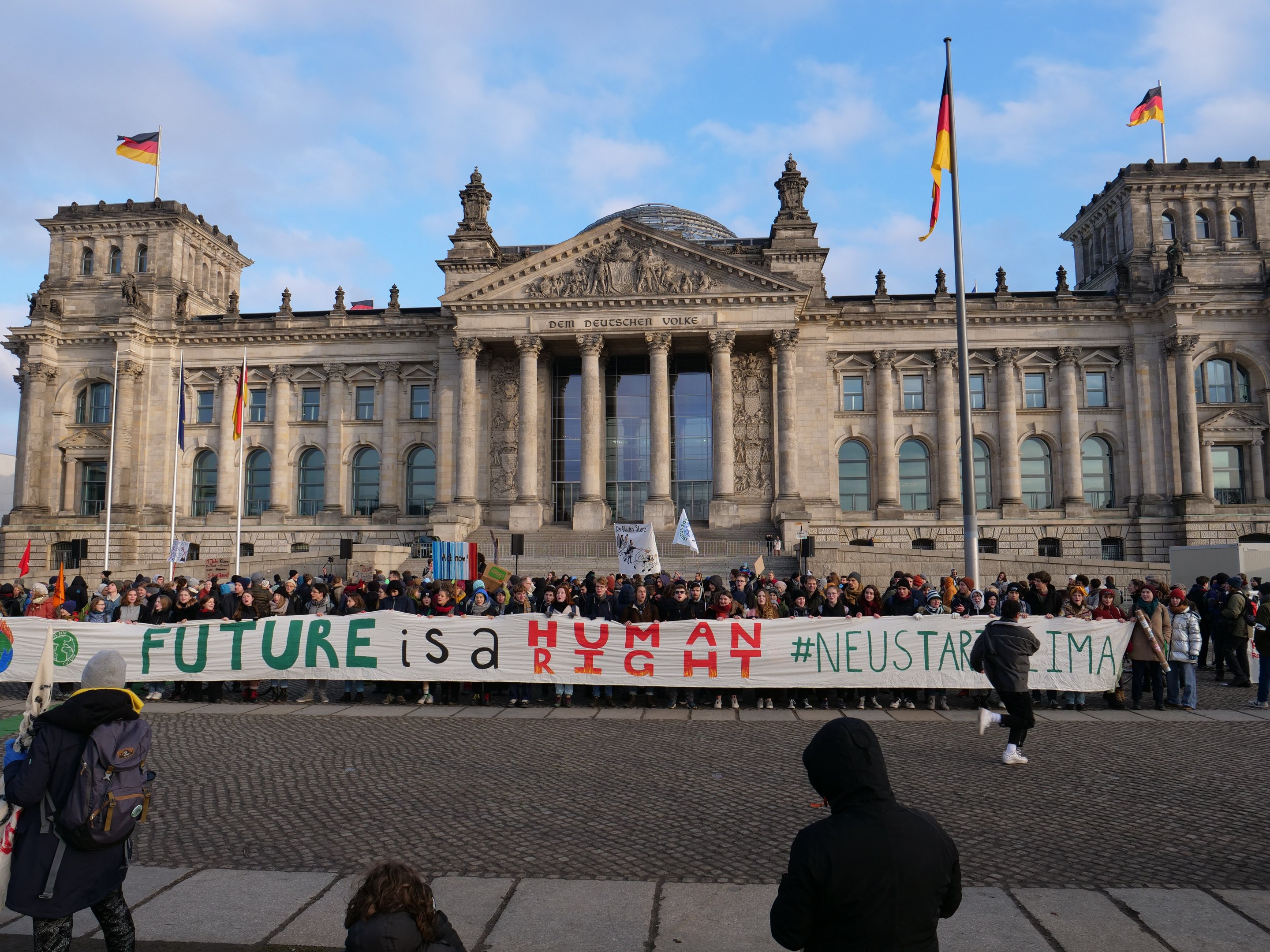 Group of people standing in front of the Reichstag building in Berlin, holding a banner with the text "Future is a Human Neustar ima" and surrounded by flagpoles.