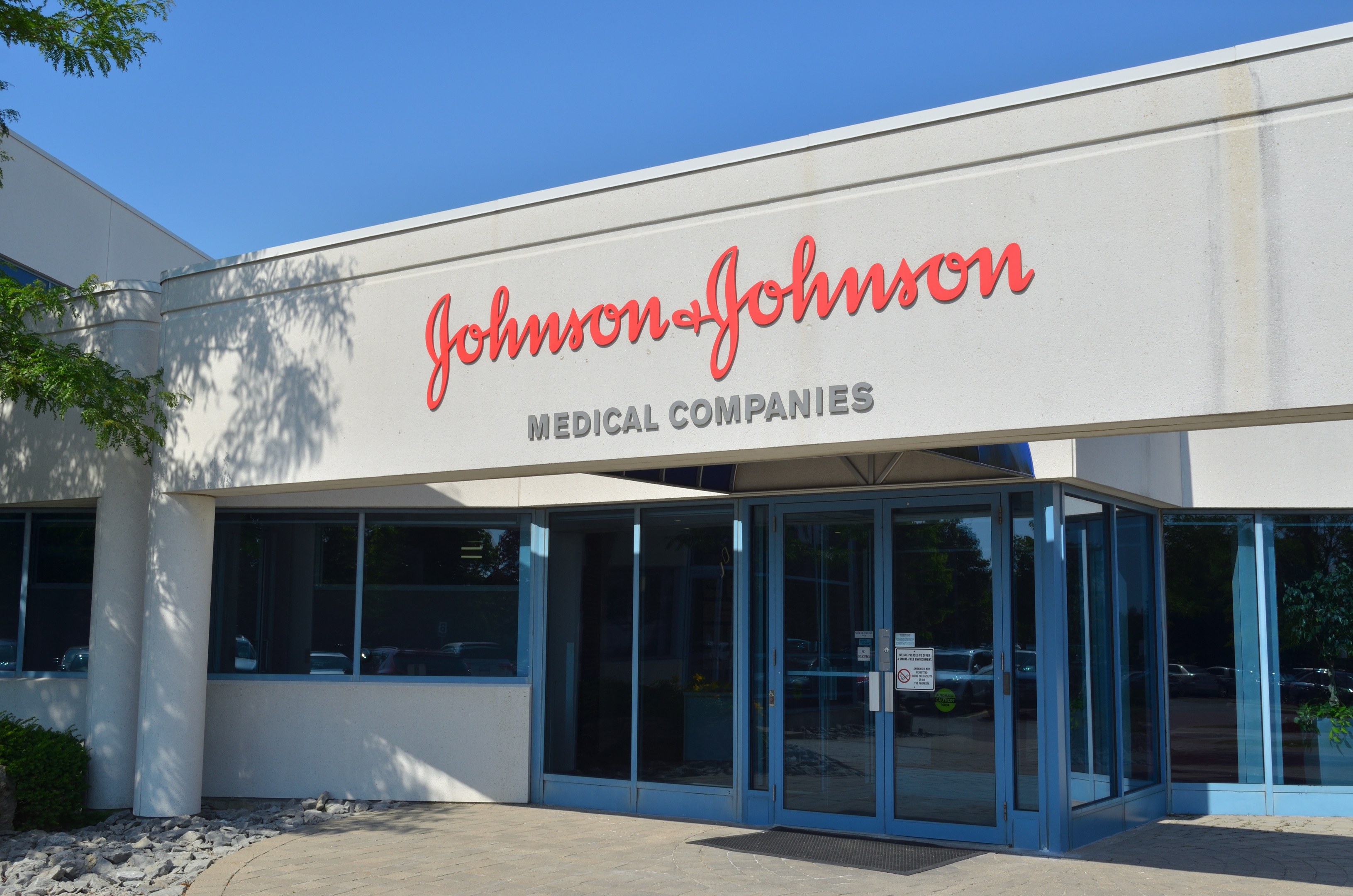 Außenansicht des Johnson & Johnson Medical Companies-Gebäudes mit Glaswänden, Text an der Fassade, Landschaftsgestaltung einschließlich Steinen und Pflanzen sowie einem Baum auf der linken Seite bei klarem blauem Himmel.