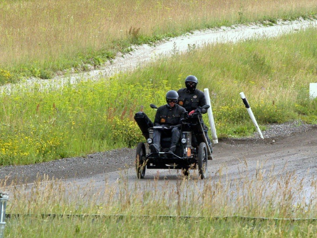 Zwei Polizisten in schwarzer Uniform und Helm fahren auf einem dreirädrigen Motorrad über einen von Gras und Pflanzen gesäumten Schotterweg mit weißen Pfosten im Hintergrund.