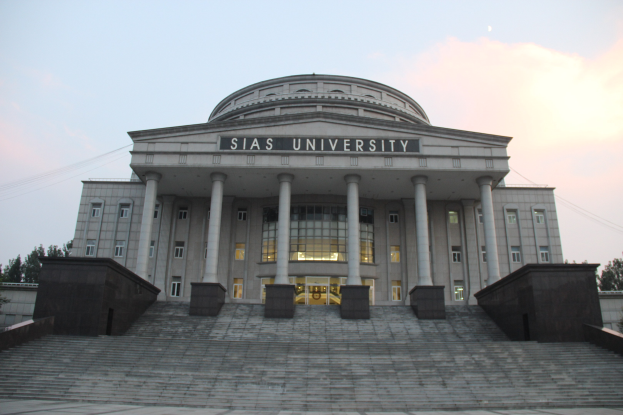Large building with pillars, windows, doors, and steps, surrounded by trees under a clear blue sky, with a university name on the facade.