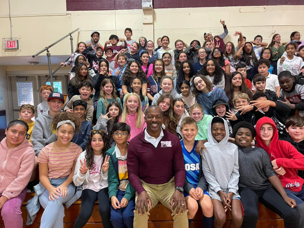 Gruppe von Schülern, die für ein Foto in einer Turnhalle posieren, einige sitzen auf der Treppe und andere stehen, eine Tür und ein Ausgangsschild sind links zu sehen.