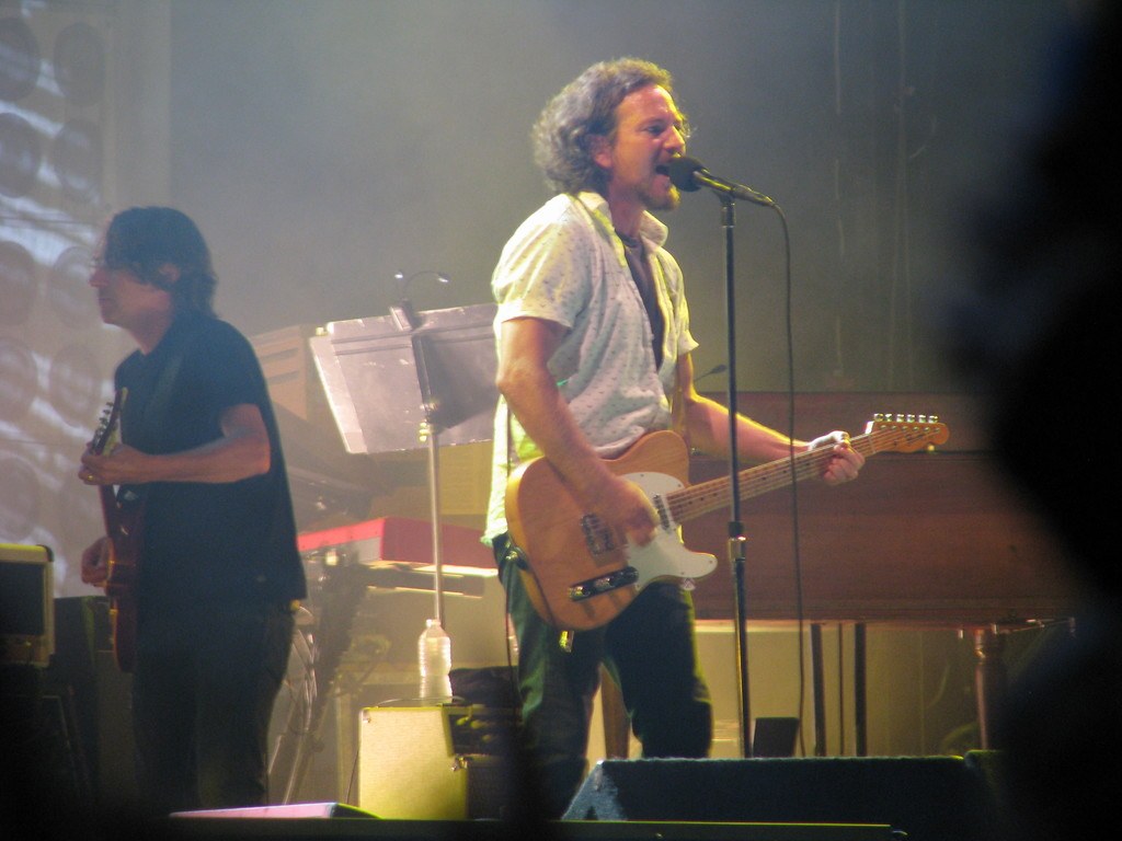 A man in a white shirt sings into a microphone while playing guitar on stage, accompanied by another man in a black shirt playing guitar, with a piano, cables, speakers, and lights visible in the background.