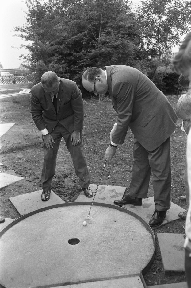 Zwei Männer in Schwarz und Weiß spielen Minigolf auf einem von Bäumen und einem Zaun umgebenen Kurs, jeder hält einen Golfschläger.