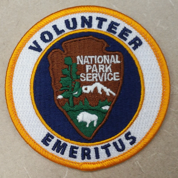 Runder Flicken mit blauem Rand und weißem Hintergrund, der mittig den schwarzen Text "Volunteer National Park Service Emeritus" in einem gelben Kreis zeigt.