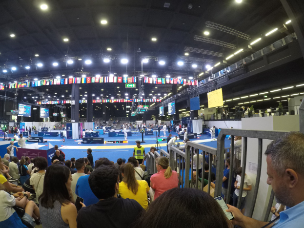 Eine große Menge Menschen sitzt und steht in einer Indoor-Sportarena mit Bildschirmen, Flaggen und Deckenbeleuchtung.