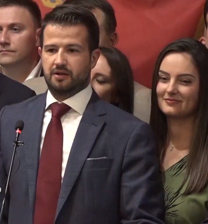 Ein Mann im Anzug spricht in ein Mikrofon an einem Rednerpult, mit einem Banner im Hintergrund und einer sichtbaren Menge, wahrscheinlich zu einem sozialen Medienverbot der Regierung.