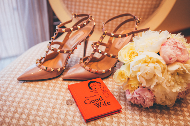 Ein Tisch zeigt ein Buch mit dem Titel "Wie werde ich eine gute Ehefrau", einen Blumenstrauß und ein Paar Valentino-Schuhe, vor einem Vorhang.