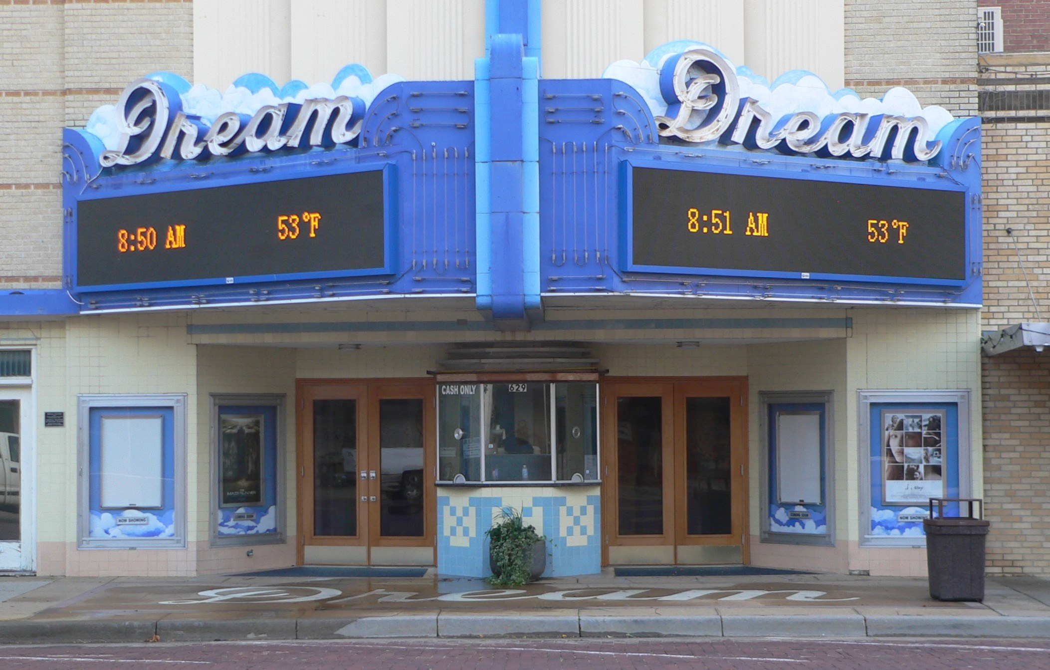 Außenansicht des Dream Theaters in St. Louis, Missouri, mit Glastüren und -fenstern, einer Schautafel, einem Mülleimer, einem Topf mit einer Pflanze und einer Straße unten.