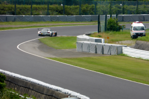 Ein Rennwagen f√§hrt auf einer Rennstrecke mit einem Polizeiwagen im Hintergrund, umgeben von Gras, Pflanzen, B√§umen und Absperrungen, mit einem Fahrzeug auf der rechten Seite und Text unten.