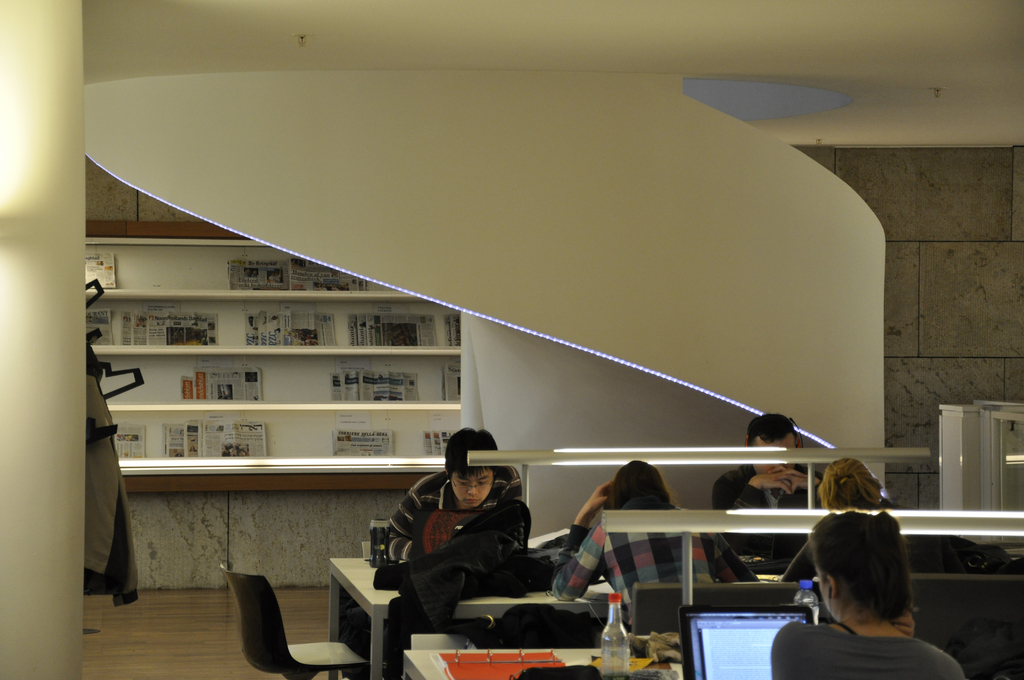 Eine Gruppe von Männern und Frauen sitzt an Tischen mit Akten, Wasserflaschen und Laptops; ein Regal mit Zeitungen ist im Hintergrund zu sehen.