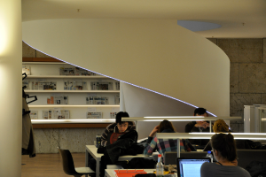 Eine Gruppe von Männern und Frauen sitzt an Tischen mit Akten, Wasserflaschen und Laptops; ein Regal mit Zeitungen ist im Hintergrund zu sehen.