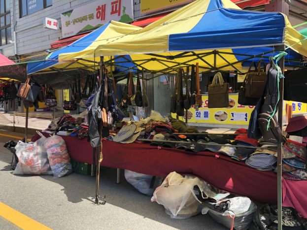 Ein Straßenmarkt mit Zelten, auf denen Taschen, Hüte und andere Gegenstände präsentiert werden, vor einem Hintergrund aus Gebäuden mit Fenstern und Texttafeln.