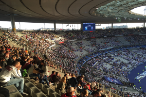 Große Zuschauermenge im Allianz Arena Stadion bei einem Fußballspiel, mit einer Bühne rechts, Fahnen, Stangen, einem Bildschirm und sichtbarem Himmel im Hintergrund.