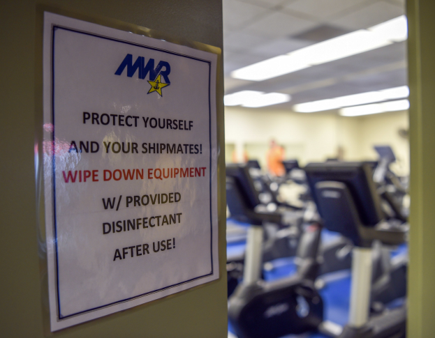 Ein Türschild mit der Aufschrift "Schützen Sie sich und Ihre Schiffskameraden" mit Laufbändern und Deckenleuchten im Hintergrund, das eine leicht unscharfe, bewegungähnliche Wirkung zeigt.