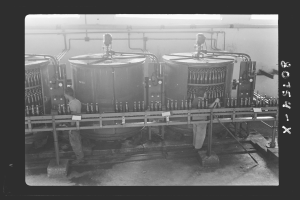 Zwei Männer in einer Fabrik, die an einer Abfüllmaschine arbeiten, mit Rohren an der Wand im Hintergrund.