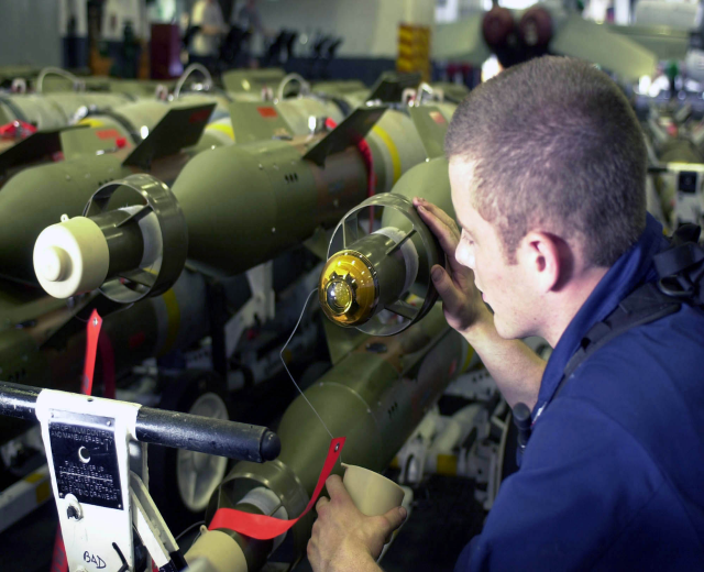 Ein Mann in einem blauen Hemd arbeitet an einer Maschine in einer Fabrik, umgeben von Raketen, mit unscharfen Menschen, einer Wand und einem Fenster im Hintergrund.