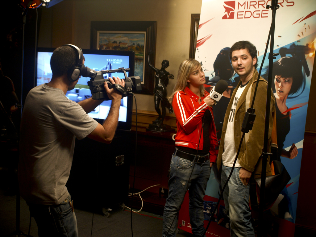 Ein Mann mit einer Videokamera und Headset filmt ein Interview zwischen einem Mann und einer Frau, mit einem Fernseher und einem gerahmten Foto im Hintergrund.