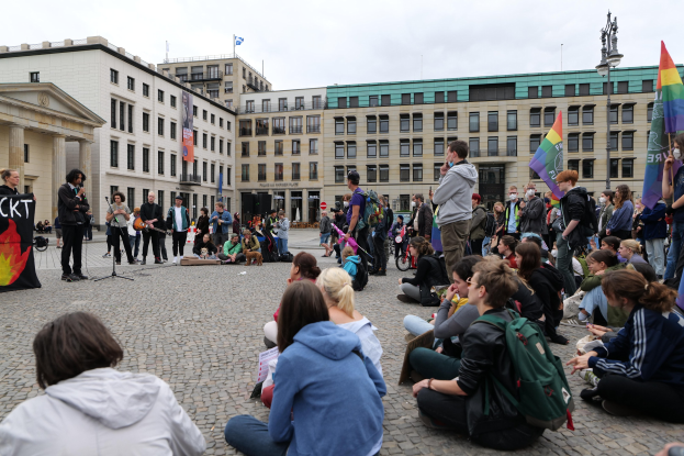 Eine Gruppe von Menschen auf dem Boden sitzend, die einer Menge mit Fahnen, Spruchbändern und einem Mikrofon auf einem Ständer gegenüberstehen, mit einer Statue, Gebäuden und einem bewölktem Himmel im Hintergrund bei einer Homo-Hass-Demonstration in Berlin, Deutschland.