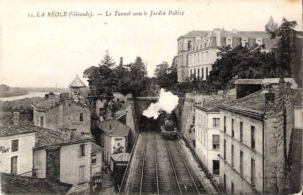 Black and white photograph of a train on tracks passing through an urban area with buildings, trees, and water, with text at the top reading "la réole gironde - le tunnel sous le jardin public".