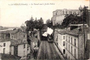 Black and white photograph of a train on tracks passing through an urban area with buildings, trees, and water, with text at the top reading "la réole gironde - le tunnel sous le jardin public".