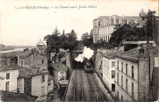 Black and white photograph of a train on tracks passing through an urban area with buildings, trees, and water, with text at the top reading "la réole gironde - le tunnel sous le jardin public".