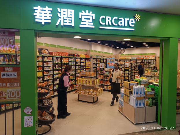 Eine Gruppe von Menschen steht vor einem Geschäft mit einem "crcare"-Schild, mit Regalen voller Lebensmittel und Flaschen im Inneren und einer Tafel mit Text auf der linken Seite.