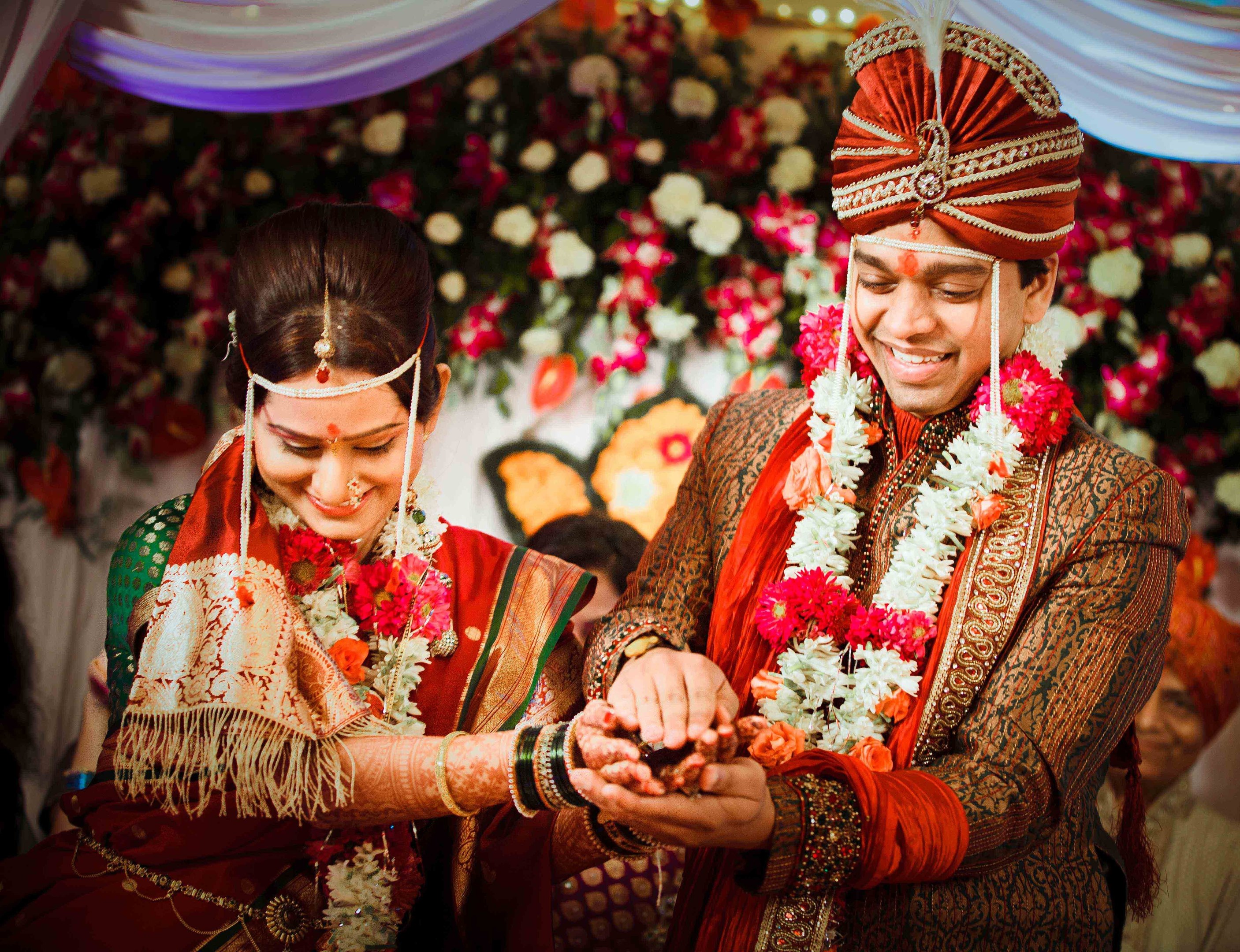 A man and woman in traditional Indian wedding attire exchange rings as guests and floral decorations are visible in the background beneath a curtain.