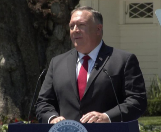 Secretary of State Mike Pompeo stands at a podium with two microphones, delivering a speech in front of a tree and plants, with a building in the background; a board with text is visible on the podium.
