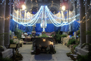 Eine Krippe in einer Kirche, geschm√√∂ckt mit Weihnachtslichtern und Deko, mit Statuen, Pflanzen, S√§ulen und einer Tafel mit Text im Vordergrund und einem unscharfen Hintergrund mit einer beleuchteten Decke.